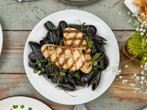 A plate of grilled mussels topped with two slices of grilled bread, garnished with herbs on a rustic wooden table.