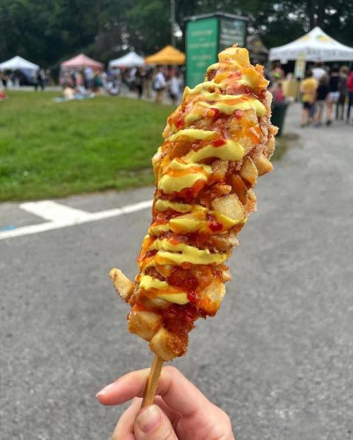 A hand holding a corn dog topped with diced potatoes, ketchup, and mustard, set against a festival background.