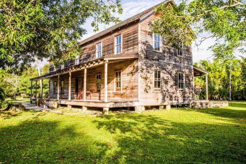 A rustic wooden house with a porch, surrounded by lush greenery and trees under a clear blue sky.