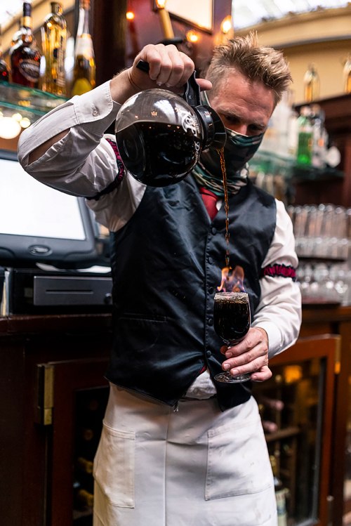 A bartender in a vest pours coffee into a glass, creating a flame on top, in a cozy bar setting.