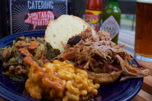A plate featuring pulled pork on a bun, macaroni and cheese, and collard greens, with sauces and a drink in the background.