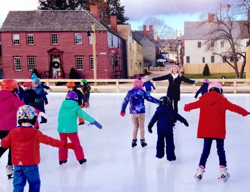 A group of children ice skating on a rink, with a woman in a black coat encouraging them in a winter setting.