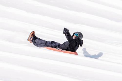 A person in winter gear sleds down a snowy slope, smiling and pointing with excitement.