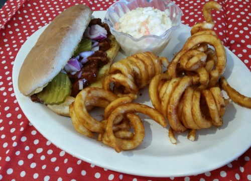 A plate with a BBQ sandwich topped with pickles and onions, curly fries, and a side of coleslaw on a polka dot tablecloth.