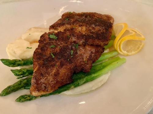 A plate featuring seasoned fish fillet on a bed of mashed potatoes and asparagus, garnished with lemon slices.