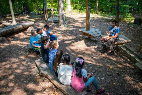 A group of children sits on logs in a forest, listening to an instructor seated nearby. Trees surround the area.