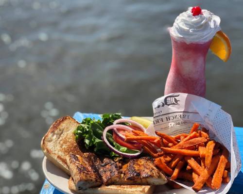 A plate of grilled fish with lettuce, onion, toast, and sweet potato fries, alongside a colorful drink topped with whipped cream.