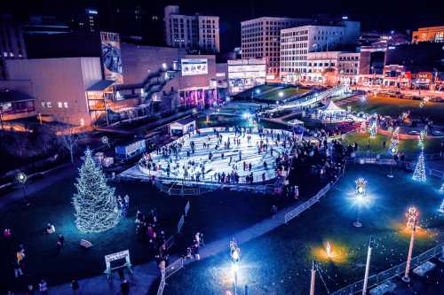 Aerial view of a bustling ice skating rink at night, surrounded by festive lights and a decorated Christmas tree.