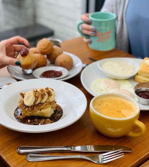 A table set with a plate of banana dessert, coffee, and a person holding a mug, with pastries in the background.
