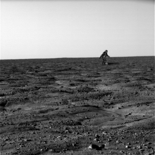 A figure in a spacesuit walks across a barren, rocky landscape under a clear sky, resembling a distant planet's surface.