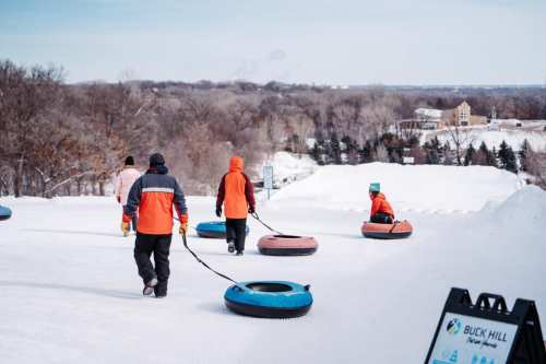 People in winter gear pull inflatable tubes up a snowy hill at a tubing park, with trees and buildings in the background.