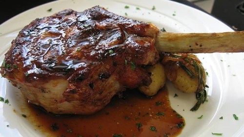 A succulent roasted meat chop with a bone, served with golden potatoes and a rich sauce, garnished with herbs.