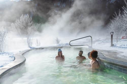 Four people relax in a steaming hot spring surrounded by snowy landscapes and misty air.