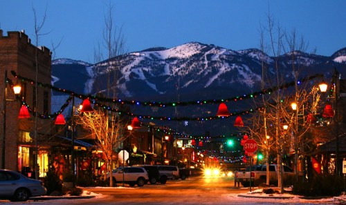 A snowy mountain backdrop with a festive street decorated with lights and holiday bells at dusk.