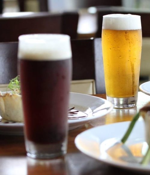 Two glasses of beer, one dark and one light, sit on a table with plates of food in the background.