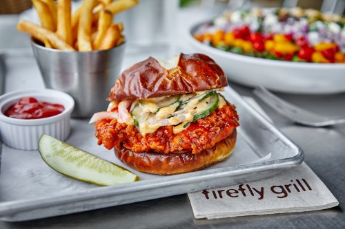 A crispy chicken sandwich on a pretzel bun, served with fries, pickles, and dipping sauce on a metal tray.
