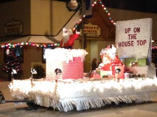 A festive parade float featuring Santa, a chimney, and colorful presents, decorated with lights and holiday cheer.
