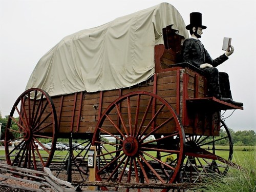 A large wooden wagon with a figure in a top hat reading a book, set against a cloudy sky.