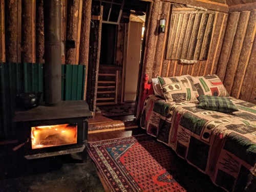 Cozy cabin interior with a wood stove, a bed covered in a patterned quilt, and rustic wooden walls.