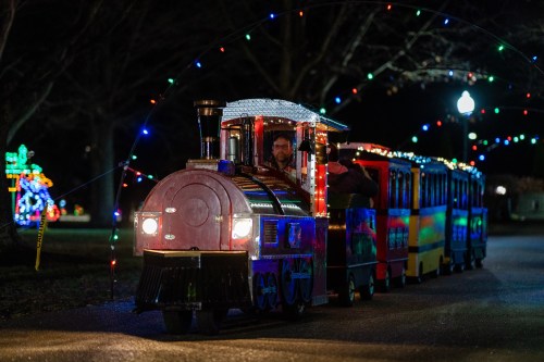 A festive train decorated with lights travels along a path, surrounded by holiday decorations in a park at night.