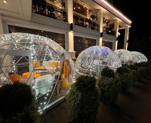 Three illuminated igloo-style dining pods lined up outside a restaurant, surrounded by greenery at night.