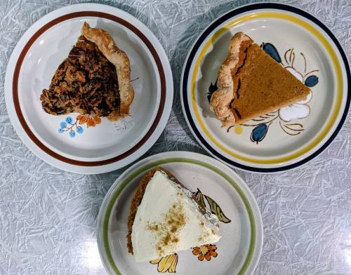 Three slices of pie on decorative plates: pecan, pumpkin, and cream pie, arranged on a textured table surface.