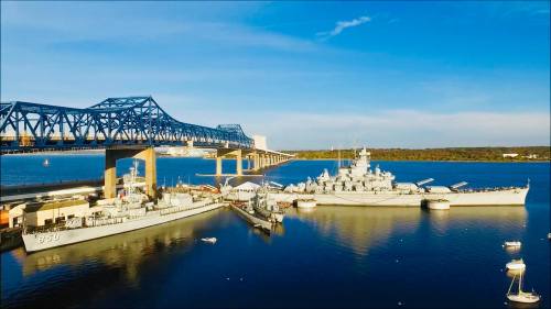 Aerial view of naval ships docked near a blue bridge over calm waters under a clear sky.