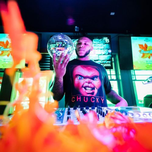 A DJ wearing a Chucky t-shirt holds a disco ball, surrounded by colorful lights and decorations.