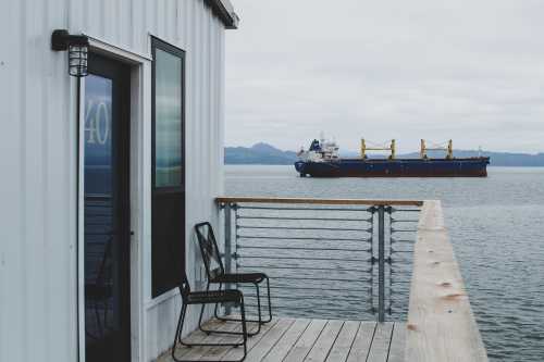 A ship sails in calm waters near a dock with a chair and a building in the foreground. Overcast sky.