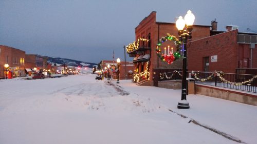 Snow-covered street decorated with holiday lights and wreaths, with buildings and lampposts in a quiet, wintry scene.