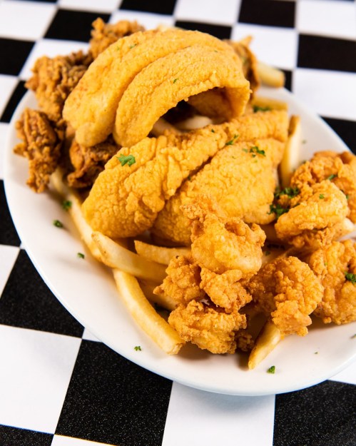 A plate of fried fish and chicken served with French fries on a checkered tablecloth.