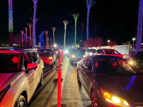 Cars lined up at night, illuminated by colorful lights on palm trees, creating a festive atmosphere.