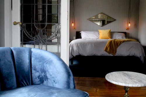 A cozy bedroom with a bed, decorative mirror, and warm lighting, viewed from a stylish blue chair and marble table.