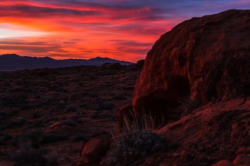 Vibrant sunset over rocky terrain, with silhouetted mountains in the background and colorful clouds in the sky.
