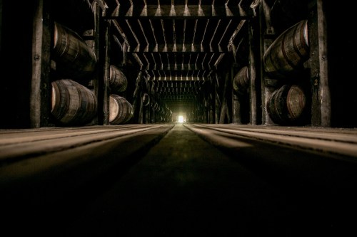 A dimly lit warehouse with wooden barrels lined along the walls, leading to a bright light at the end of the corridor.