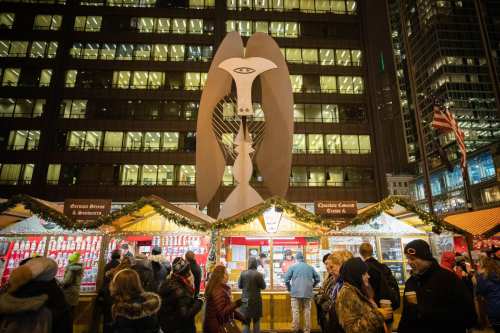 A large abstract sculpture stands over festive market stalls, with people enjoying the holiday atmosphere in a city setting.