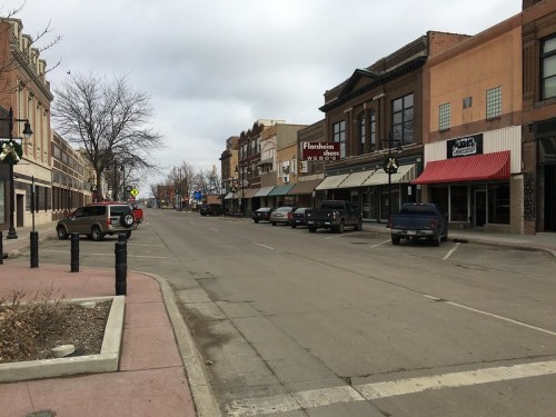 A quiet downtown street lined with shops and parked cars under a cloudy sky.