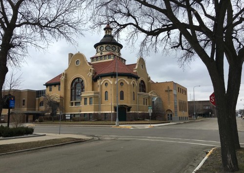 Historic building with a distinctive dome, surrounded by trees and a quiet street under a cloudy sky.