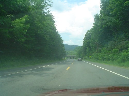 A scenic road surrounded by lush green trees, leading towards a bridge in the distance under a partly cloudy sky.
