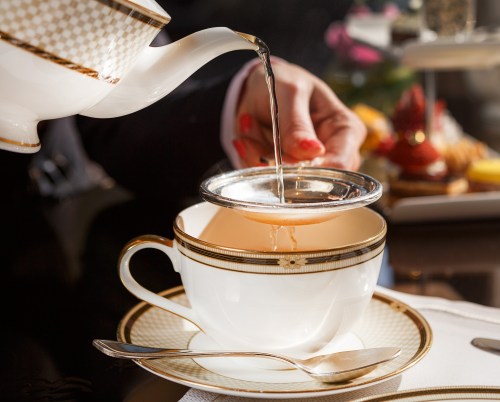 A hand pours tea through a strainer into an elegant cup with a gold rim, set on a table with a spoon nearby.