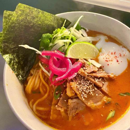 A bowl of ramen with sliced meat, soft egg, lime, pickled onions, greens, and seaweed on top.