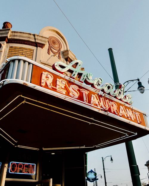 Neon sign of the Arcade Restaurant, featuring vintage lettering and a bright "OPEN" sign, against a clear sky.