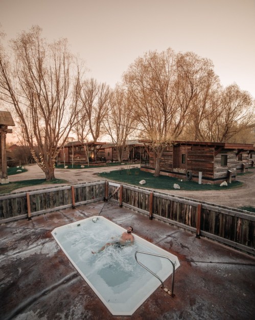A person relaxes in a hot tub surrounded by trees and rustic cabins at sunset.
