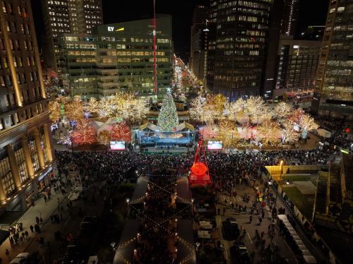 A vibrant city square at night, adorned with festive lights and a large Christmas tree, bustling with a crowd.