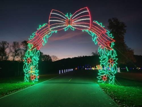Colorful holiday light arch with a bow, framing a dark road at dusk, surrounded by trees and festive decorations.