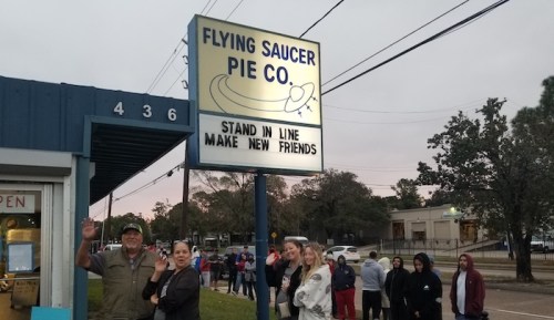 A line of people waiting outside Flying Saucer Pie Co., with a sign that reads "Stand in line, make new friends."