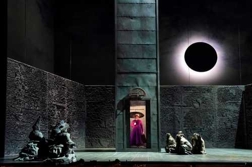 A dramatic stage scene featuring a figure in a purple robe, surrounded by dark sculptures and a large black circle overhead.