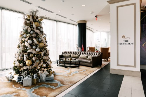 A decorated Christmas tree stands beside a cozy seating area in a modern lobby with large windows.