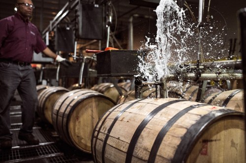 A worker operates machinery as water splashes over wooden barrels in a distillery.