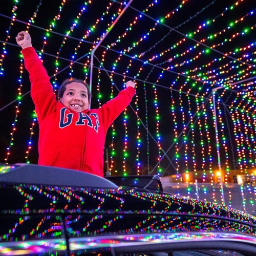 A joyful child in a red hoodie raises their arms in excitement under colorful holiday lights.